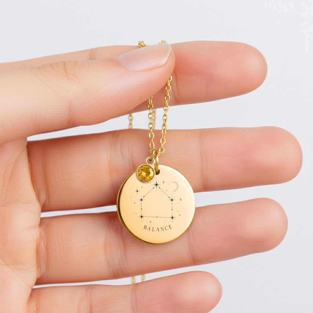 Collier constellation Balance tenu entre les doigts, médaille ronde gravée et pierre de naissance